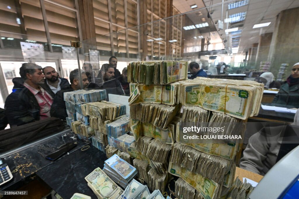 CORRECTION / People queue in front of a teller next to a pile of banknotes of different currencies at a commercial bank affiliated with Syria's central bank in Damascus on December 16, 2024. Governments worldwide are stepping up efforts to engage with Syria's new interim rulers, just over a week after Islamist-led rebels ousted president Bashar al-Assad, ending decades of brutal rule and civil war. (Photo by Louai Beshara / AFP) / "The erroneous CAPTION appearing in the metadata of this photo by Louai Beshara has been modified in AFP systems. Please immediately remove the erroneous mention[s] from all your online services and delete it (them) from your servers. If you have been authorized by AFP to distribute it (them) to third parties, please ensure that the same actions are carried out by them. Failure to promptly comply with these instructions will entail liability on your part for any continued or post notification usage. Therefore we thank you very much for all your attention and prompt action. We are sorry for the inconvenience this notification may cause and remain at your disposal for any further information you may require." (Photo by LOUAI BESHARA/AFP via Getty Images)